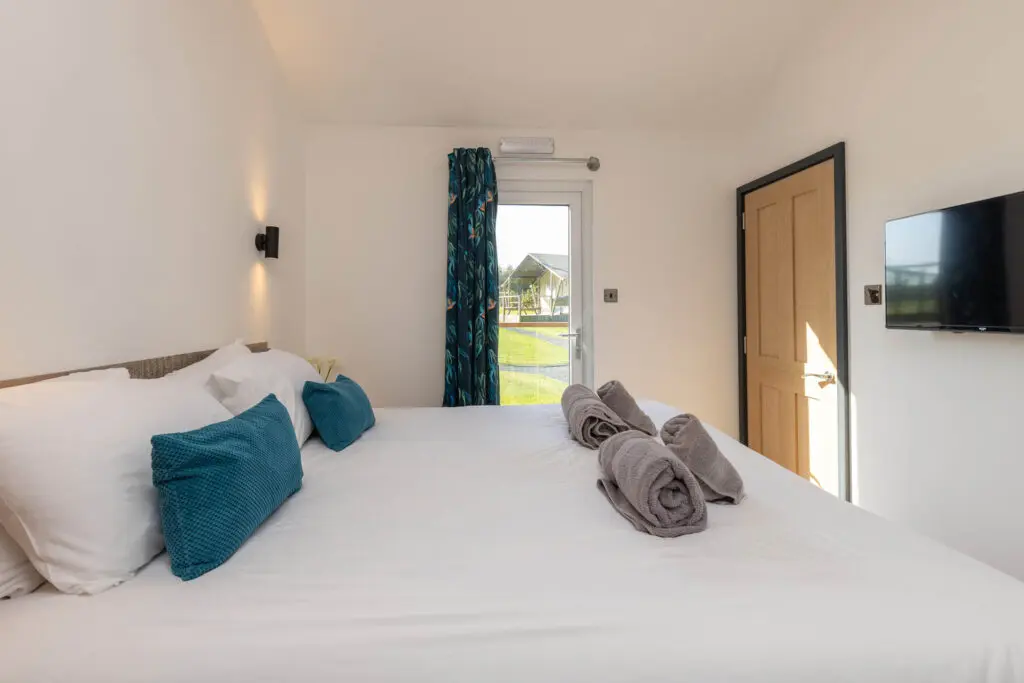 Double bed with teal upholstered headboard inside holiday lodge accommodation at Little Pig Wellness, looking out over green countryside near Bude, North Cornwall