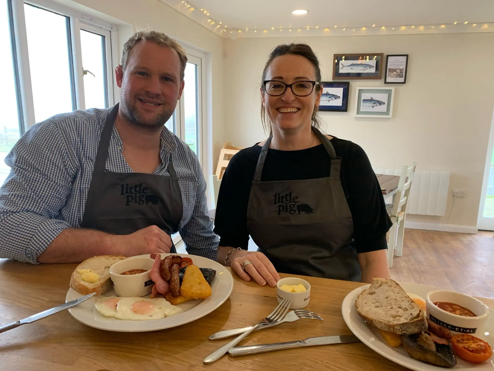 Emma and James, founders of Little Pig Wellness, wearing Little Pig branded aprons in the café near Bude, Cornwall